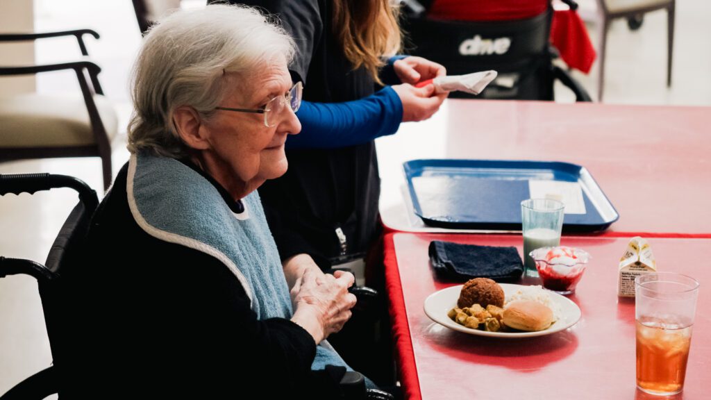 A woman sits down to eat dinner at a skilled nursing facility nursing home